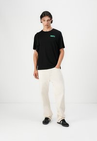 Camiseta de algodón negra con texto verde, combinada con pantalones de color crema. Modelo con zapatillas negras, de pie contra un fondo blanco.