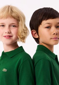 Gröna polo skjortor i texturerad tyg, med krage och broderad logotyp, som bärs av två barn som står rygg mot rygg.