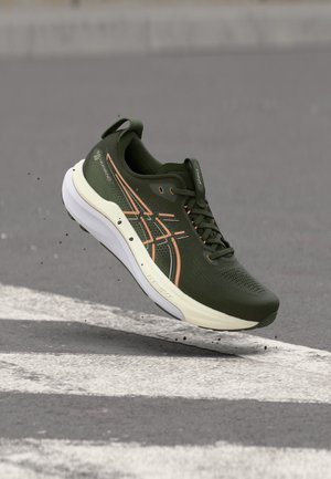 Chaussure de course vert foncé et beige en l'air au-dessus d'une surface asphaltée avec des marquages routiers blancs et de petits débris.