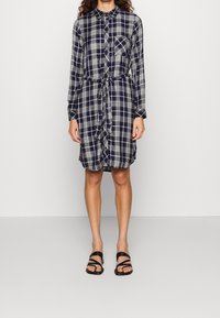 Abito a camicia a quadri blu navy e bianco con maniche lunghe, chiusura frontale con bottoni, taschino sul petto e dettaglio in vita annodato. Abbinato a sandali neri con cinturini.