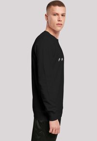 Svarte langermede sweatshirt med rund hals og ribbede mansjetter, med hvit tekst "F.R." på brystet, iført med blanke, svarte bukser.