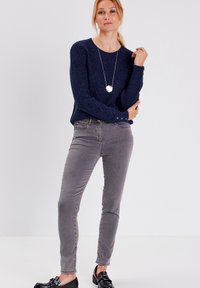 Marineblauer Strickpullover mit strukturiertem Muster, graue Slim-Fit-Hosen und schwarze Schuhe. Eine Halskette mit einem runden Anhänger vervollständigt den Look.