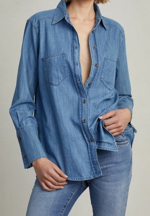 Personne portant une chemise en denim bleu clair partiellement boutonnée avec poches et un jean assorti sur un fond neutre.