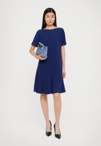 Femeie îmbrăcată într-o rochie bleumarin de lungime până la genunchi și cu tocuri negre, ținând o poșetă mică albastră, pe un fond alb.