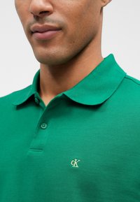 Zelená polo tričko vyrobené z textúrovaného materiálu, s golierom, troma gombíkmi a malým zlatým logom CK na ľavej hrudi.