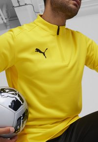 Giacca sportiva gialla con design a mezza zip e logo Puma nero. Tessuto morbido con pannelli laterali in rete. Tiene un pallone da calcio argento e nero.