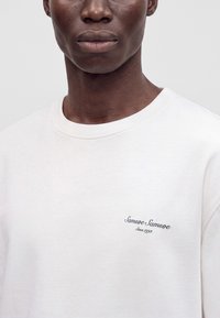 Personne à la peau foncée portant un sweat-shirt blanc à col rond avec un petit logo texte sur le côté gauche de la poitrine indiquant "Samsøe Samsøe Since 1993".