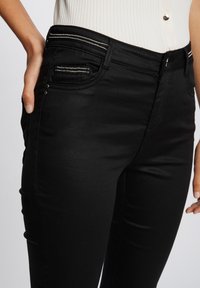 Jeans negros con una tela suave y elástica. Presenta un bolsillo frontal con un cierre metálico y costura decorativa.