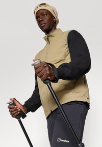 Gilet beige e nero con zip e tasche laterali, indossato sopra una maglietta nera a maniche lunghe. La persona tiene dei bastoncini da trekking con impugnature grigie.