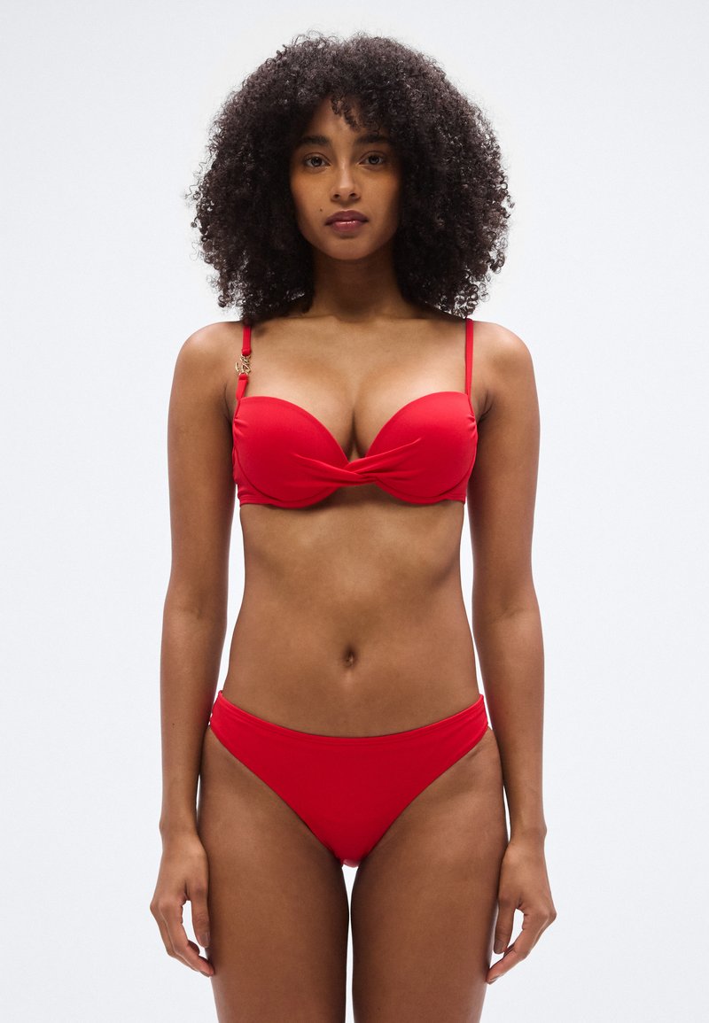 Κόκκινο μπικίνι με στριφτό μπροστινό σχέδιο, λεπτές τιράντες και χρυσές μεταλλικές λεπτομέρειες. Λεία υφή και εφαρμοστή γραμμή, παρέχοντας μέτρια κάλυψη.