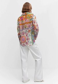 Chemise colorée à motifs présentant des designs floraux et géométriques, avec une coupe décontractée, des manches longues et un pantalon large blanc.