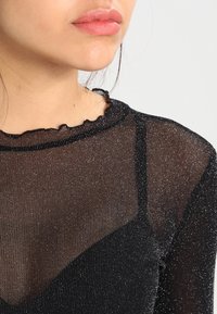 Svart genomskinlig topp med glittrig textur, med en scalloped halsringning och tunna axelband, lager över en solid svart inre del.