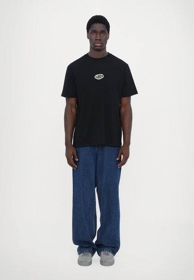 Camiseta de algodón negra con un logo en el pecho, combinada con jeans de mezclilla azul de pierna ancha y zapatillas grises, exhibida contra un fondo plano.