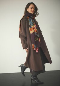 Lang brun frakke med en blød tekstur, parret med et flerfarvet blomster tørklæde. Sorte spidse hæle støvler fuldender outfittet.