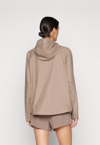 Veste capuchonnée de couleur brun clair en matériau lisse, avec des poignets élastiques et une coupe décontractée. Vue de dos, portée avec un short assorti.