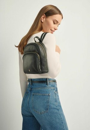 Młoda kobieta ubrana w biały prążkowany sweter i niebieskie dżinsy, pozująca z małym czarnym skórzanym plecakiem narzuconym na jedno ramię.