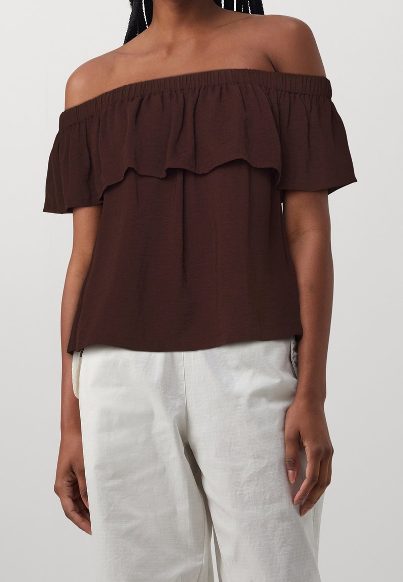 Femme portant un haut marron foncé avec épaules dénudées et volants, ainsi qu'un pantalon blanc ample sur un fond clair uni.