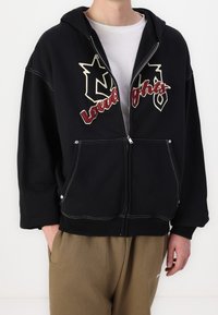 Sweat à capuche noir à fermeture éclair, en coton, avec des coutures contrastantes blanches et un logo graphique beige et bordeaux sur le devant.