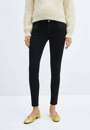 Frau trägt schwarze Slim-Fit-Jeans, cremefarbenen Zopfpullover und goldene Ballerinas, steht vor einem einfachen hellen Hintergrund.