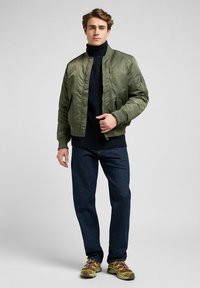 Giacca bomber verde con polsini e vita a costine, tasche con zip, abbinata a un maglione scuro, jeans blu e sneakers marroni.