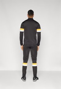 Μαύρο αθλητικό κοστούμι φτιαγμένο από ελαφρύ ύφασμα; διαθέτει πορτοκαλί και λευκές ρίγες στα μανίκια και στα παντελονιά; εφαρμοστή σχεδίαση.