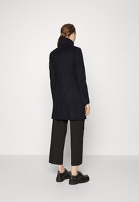 Černý vlněný kabát s vysokým límcem, přizpůsobený design a úzký střih, doplněný širokými černými kalhotami a robustními černými botami.