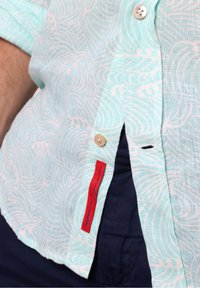Chemise bleu clair à manches courtes avec un motif de vagues blanches, un col boutonné et un détail d'étiquette rouge sur la couture latérale.