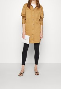 Beżowa bawełniana sukienka koszulowa z guzikiem z przodu i dwiema kieszeniami na piersi, w połączeniu z czarnymi legginsami i czarnymi klapkami, trzymająca białą kopertówkę.