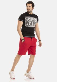 Zwarte T-shirt met grafische print, gecombineerd met rode shorts, voorzien van zijzakken en een trekkoord. G gedragen met witte en rode sportschoenen.