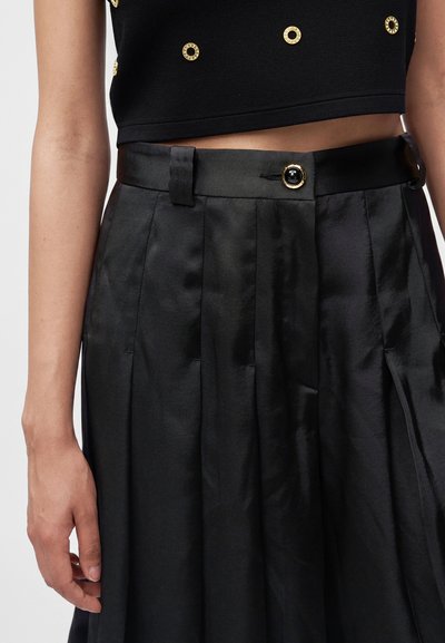 Femme portant une jupe plissée satin noir taille haute avec un bouton doré et noir, associée à un crop top noir orné de clous circulaires dorés.