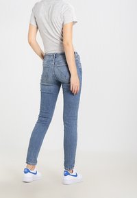 Vaqueros de mezclilla azul ajustados con un lavado desgastado, diseño de cinco bolsillos y costura en el bolsillo trasero. Combinados con zapatillas blancas con detalles en azul.