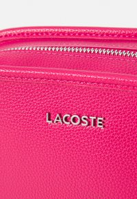Rožinė tekstūruota odinė rankinė su sidabriniu užtrauktuku ir Lacoste logotipu. Pasižymi lygiu paviršiumi ir geometriniu raštu medžiagoje.