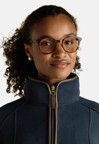 Mladá žena s kučeravými vlasmi, nosiaca okuliare s korytnačkovým vzorom a zlaté kruhové náušnice, v námorníckej flísovej bunde so zipsom a vysokým golierom.