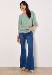 Mintgrüne, mehrlagige Bluse mit kurzen Ärmeln und gewelltem Saum, kombiniert mit dunkelblauen Schlagjeans und beigen High-Heels.