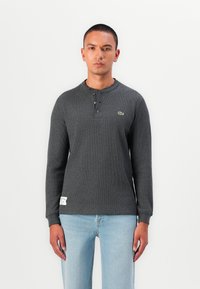 Cămașă henley cu mânecă lungă, gri, din tricot texturat, cu placket cu trei nasturi și un mic accent logo verde. Asortată cu blugi albaștri deschiși.