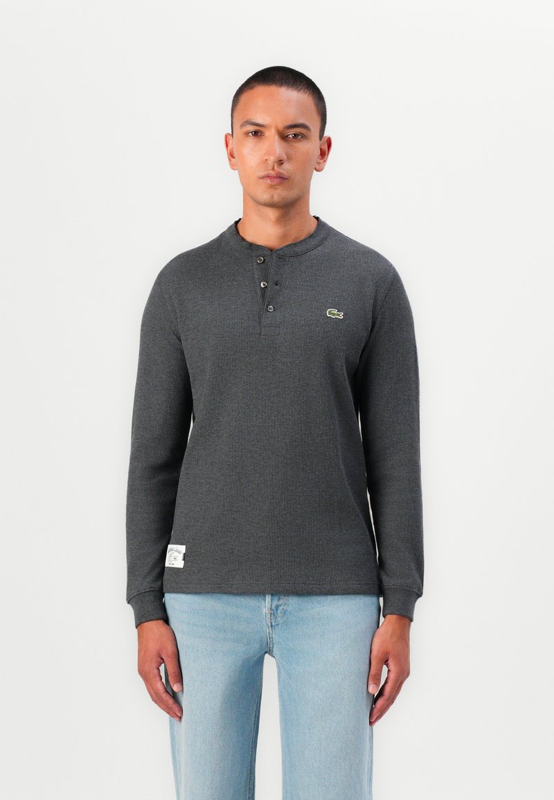 Cămașă henley cu mânecă lungă, gri, din tricot texturat, cu placket cu trei nasturi și un mic accent logo verde. Asortată cu blugi albaștri deschiși.