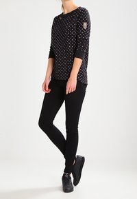 Camiseta negra de manga larga con lunares de colores y cuello redondo, combinada con pantalones negros ajustados y zapatillas negras. Detalle notable en el parche de la manga.