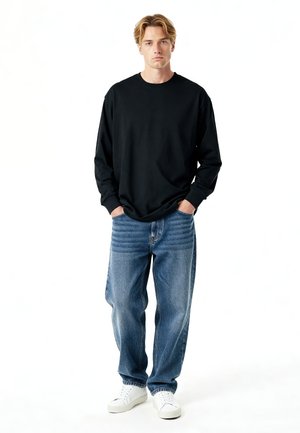 Giovane uomo in piedi con le mani nelle tasche, che indossa una maglia a maniche lunghe nera, jeans blu larghi e sneakers bianche su sfondo bianco.