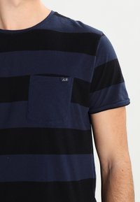 Stribet t-shirt i marineblå og sort lavet af bomuld. Har en rund halsudskæring og en brystlomme med et lille logo-tag. Korte ærmer.