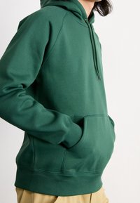 Vihreä hupullinen collegepaita pehmeästä kankaasta, jossa on etutasku, resorit hihansuissa ja kiristysnauhalla varustettu huppu. Yksinkertainen malli ilman kuvioita.