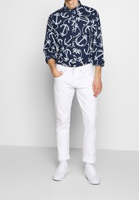 Chemise bleu marine à boutons avec un motif d'ancre blanche, assortie avec un pantalon blanc retroussé et des baskets blanches. Tissu lisse, coupe décontract�ée, quincaillerie minimale.
