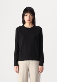 Maglione nero a maglia con maniche lunghe e colletto rotondo, caratterizzato da un piccolo logo sul petto. Abbinato a pantaloni a gamba larga di un delicato colore crema.