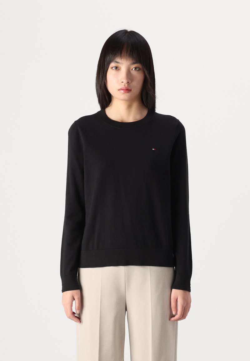 Maglione nero a maglia con maniche lunghe e colletto rotondo, caratterizzato da un piccolo logo sul petto. Abbinato a pantaloni a gamba larga di un delicato colore crema.