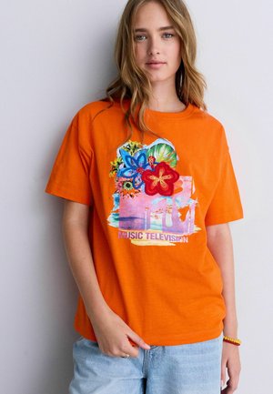 Młoda kobieta ubrana w jaskrawo pomarańczowy T-shirt z kolorowym kwiatowym logo MTV Music Television, w połączeniu z jasnoniebieskimi dżinsami i bransoletką z koralików.