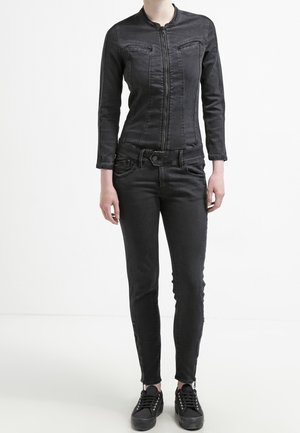 Frau trägt eine eng anliegende schwarze Jacke mit Reißverschluss, schwarze Skinny-Jeans und schwarze Turnschuhe, steht vor einem einfarbigen weißen Hintergrund.