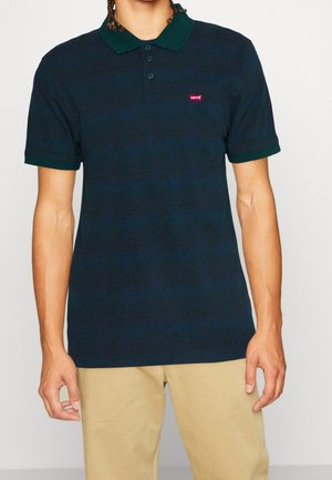 Mężczyzna ubrany w granatowo-ciemnozieloną koszulkę polo Levi's z trzema guzikami, w połączeniu z beżowymi spodniami, stoi na jednolitym tle.