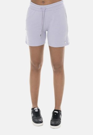 Jambes portant un short à cordon violet clair et des baskets noires à semelles blanches, debout devant un fond uni.