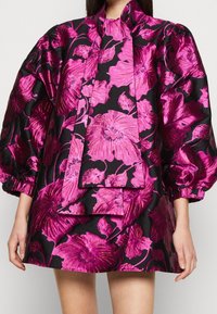 Kort, svart klänning med ett blommönster i lysande fuchsias färg. Har voluminösa ärmar och en knytbanddetalj vid kragen. Skimrande tyg.