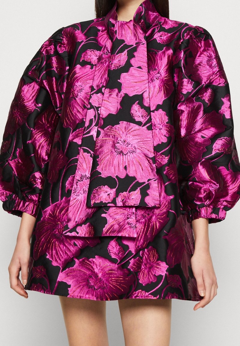 Kort, svart klänning med ett blommönster i lysande fuchsias färg. Har voluminösa ärmar och en knytbanddetalj vid kragen. Skimrande tyg.