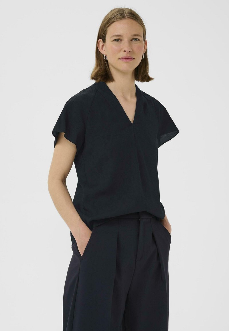 Blusa nera a maniche corte con scollatura a V, tessuto leggero, vestibilità rilassata e una texture sottile. Abbinata a pantaloni scuri a fit morbido.